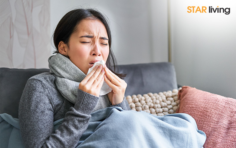 Sick woman sitting on a couch, sneezing into a tissue while wrapped in a blanket and scarf.